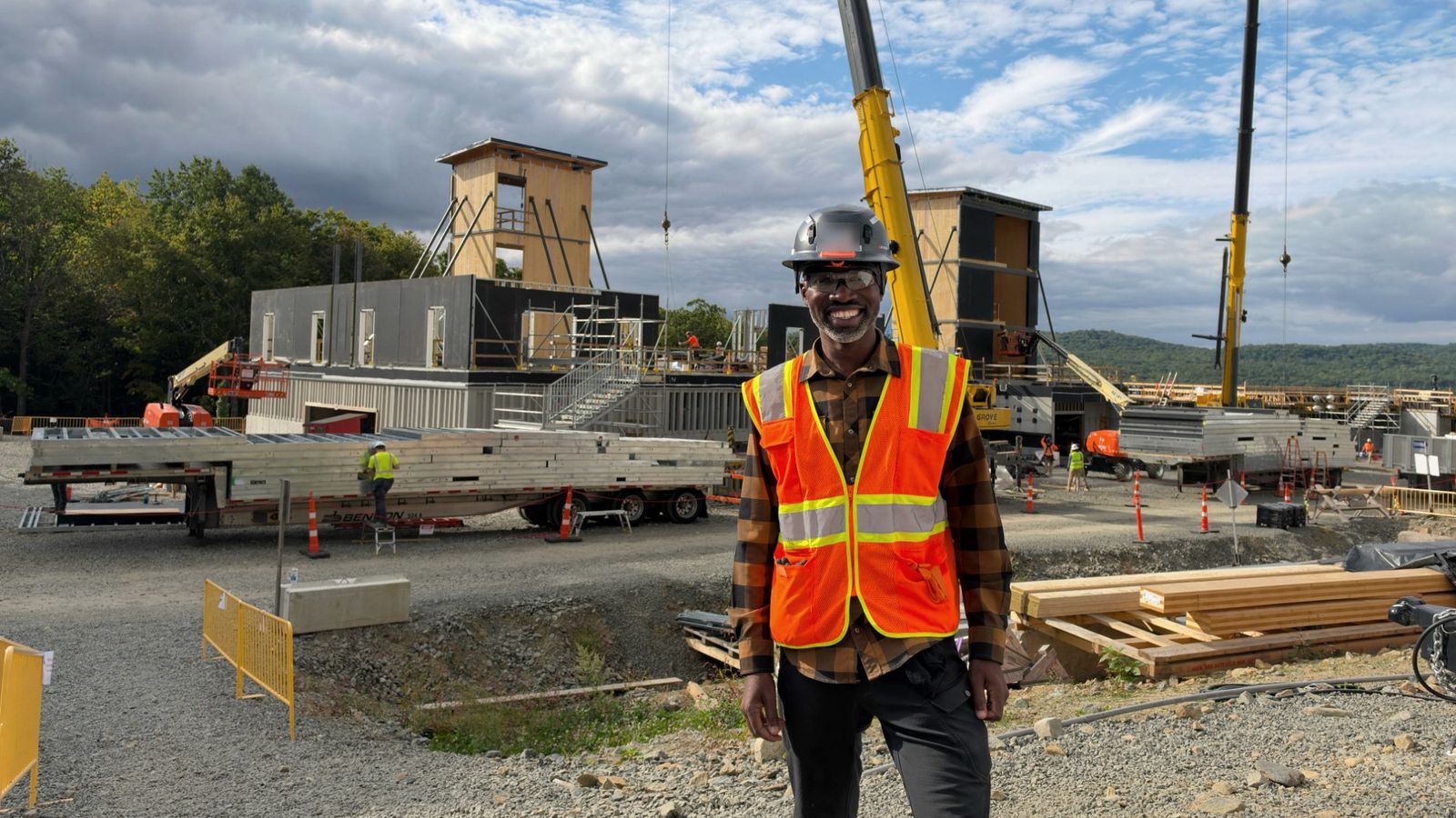 Kobe in a hard hat and safety vest, smiling on an active construction site with cranes and modular building sections behind him