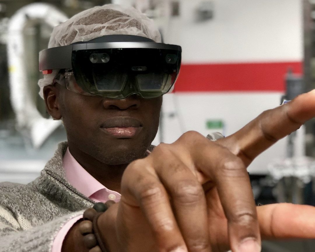 Kobe wearing a mixed-reality headset over a hairnet on a plant floor, pointing at a virtual control